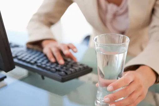 Стакан с водой стоит на столе перед человеком с компьютером